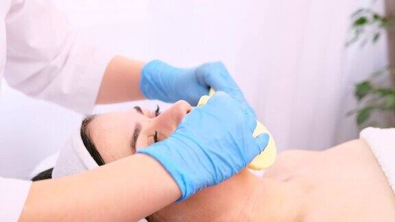 美容师正在给一位戴着医用手套的年轻女子做美容手术用两块海绵擦拭她脸上的面霜