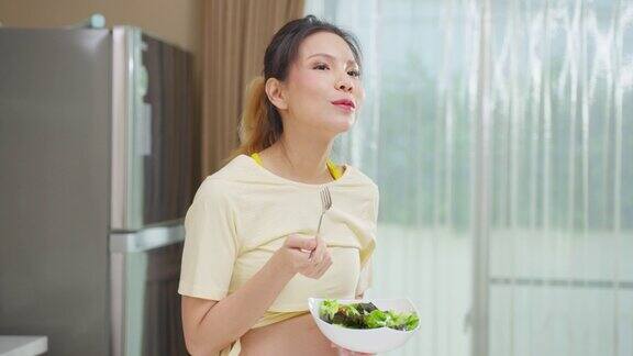 亚洲美丽的孕妇在家里的厨房吃健康的食物迷人的怀孕女孩站在厨房里心情愉快享受着在碗里吃绿色蔬菜沙拉的健康