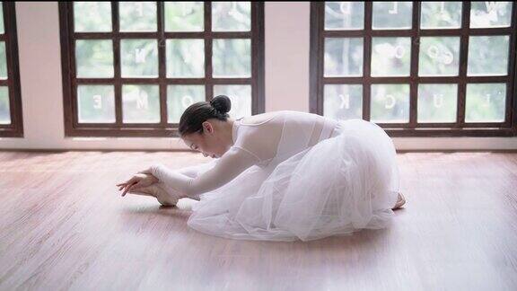 穿着白色芭蕾舞裙的年轻亚洲芭蕾舞演员坐在地板上练习芭蕾舞身体向前弯曲