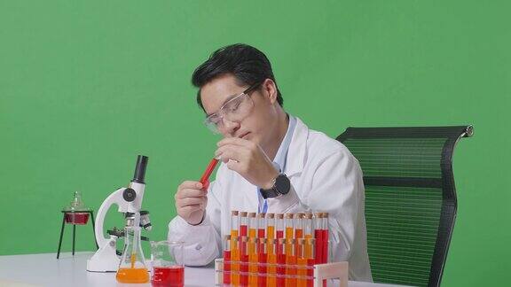 侧视图:在绿屏背景的实验室里亚洲男性科学家用显微镜看着试管里的红色液体在桌子上摇头