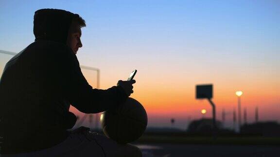黄昏时分在户外篮球场上带着篮球和智能手机的年轻人