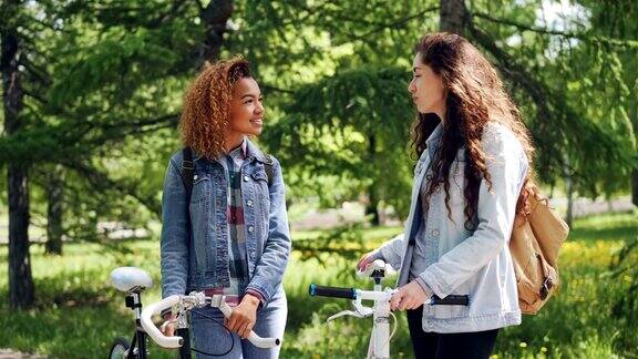 两个漂亮的女自行车手拿着自行车站在公园里聊天拍摄美丽的城市公园有许多树木和绿色的草坪