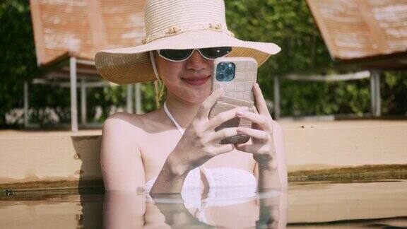手持智能手机的年轻亚洲女性正在度假酒店的室外泳池里休息