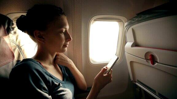 日落时坐在飞机窗口附近的女游客在飞行中使用手机