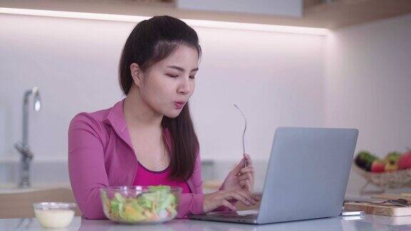 穿着运动服的年轻亚洲女性看着笔记本电脑在厨房里吃着蔬菜沙拉健康的饮食和运动的理念