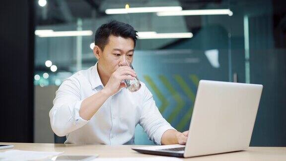 亚洲员工在现代办公室工作坐在笔记本电脑前喝着玻璃杯里的清水商人
