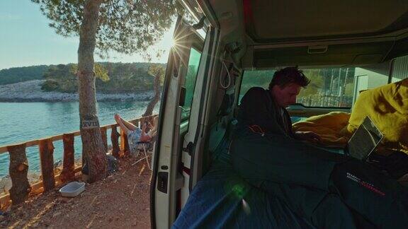 一个男人躺在他的面包车里晚上在他的笔记本电脑上工作面包车停在海边夕阳作为背景面包车生活