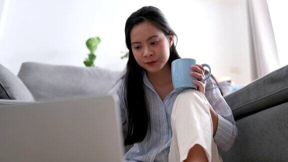 一位在客厅使用笔记本电脑的亚洲女性商业女性在笔记本电脑屏幕上思考一个新项目在家里工作的地方喝咖啡的肖像在家工作商业女性和工作理念