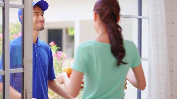 亚洲年轻的快递员在家里给女顾客送包裹穿着蓝色制服的迷人邮递员正在工作把一盒水果和蔬菜杂货送到家门口的女人面前