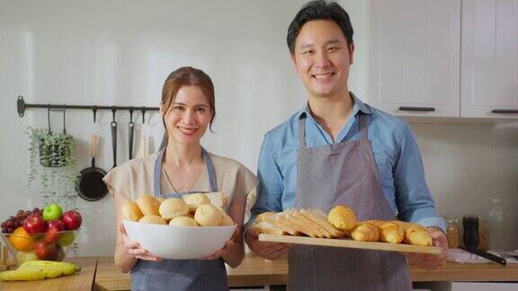 亚洲年轻夫妇拿着一碗面包看着镜头的肖像有魅力的男女系着围裙花时间在家里做饭做早餐保健饮食与健康食品理念