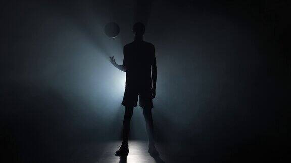 一个年轻的篮球运动员把球抛起来的剪影高大的年轻运动员在运动制服摆姿势在一个黑暗的工作室背景与烟和背光慢动作准备59.94fps