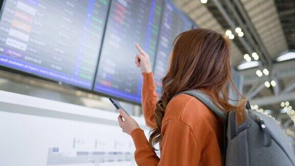 年轻的亚洲女子在国际机场用手机在航班信息板上查询航班
