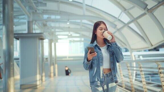 年轻的亚洲女人带着手机和咖啡杯走在城市里快乐的女人在户外用手机和热饮放松