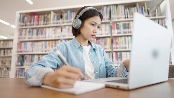 亚洲女大学生在图书馆戴着耳机在笔记本电脑上上网学习在图书馆为考试而学习