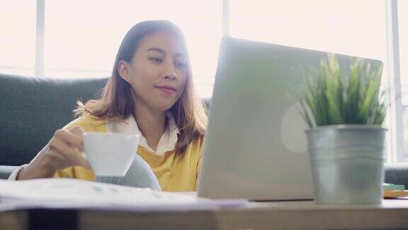 美丽的年轻微笑的亚洲女人工作在笔记本电脑和喝咖啡在家里的客厅亚洲商业女性工作文件财务和计算器在她的家庭办公室享受在家的时光