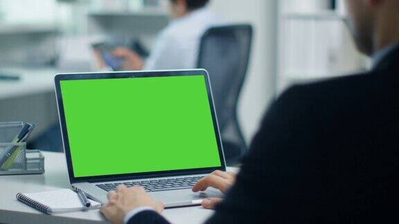 商人用绿色屏幕的笔记本电脑工作他的同事正在平板电脑上工作办公室是现代和明亮的