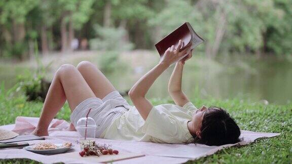 迷人的亚洲年轻女子在阳光明媚的日子里在美丽的绿色花园外野餐和读书在夏日假期放松