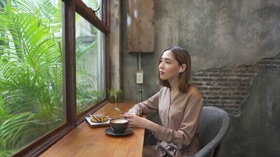 4K亚洲女人坐在窗边喝拿铁咖啡耳机里听着音乐