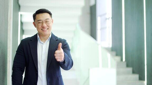 微笑的亚洲商人竖起大拇指像手势一样站在现代办公大楼里