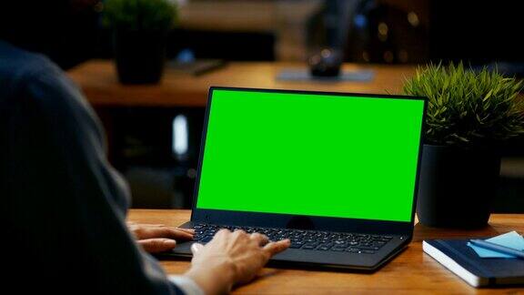女办公室职员在她的办公桌上用一台模拟绿色屏幕的笔记本电脑工作过肩镜头她坐在创意办公室的木桌前