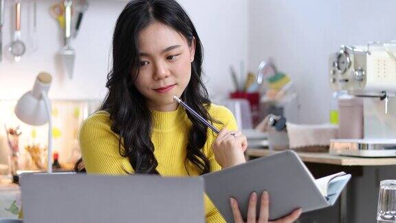 年轻的亚洲女性在家里的厨房里用笔记本电脑工作她正在网上上烹饪课并在网上写食谱笔记