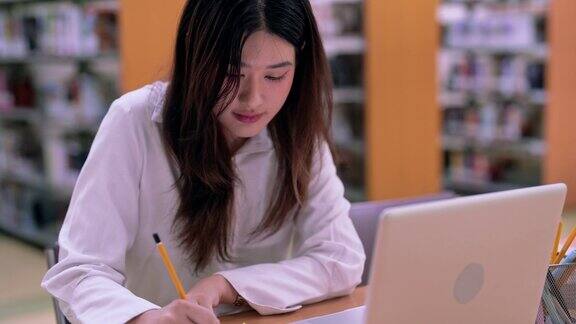 身着便服的亚洲女学生正在大学图书馆用笔记本电脑为论文做笔记