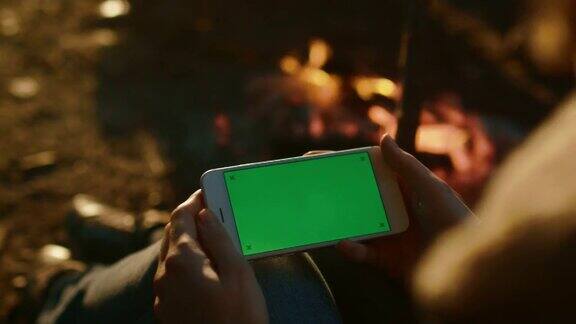 晚上女孩在篝火旁拿着一台绿色屏幕的风景模式智能手机