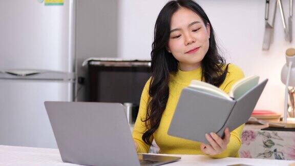 年轻的亚洲女性在家里的厨房里用笔记本电脑工作她正在网上上烹饪课并在网上写食谱笔记