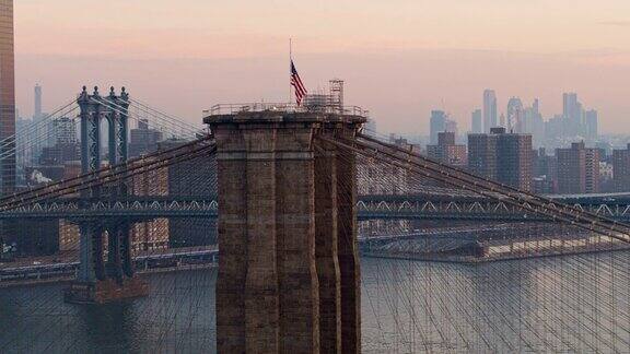 在清晨的黎明时分俯瞰布鲁克林大桥塔顶飘扬的美国国旗遥望曼哈顿无人机视频与平移摄像机运动