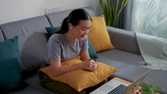 亚洲妇女戴着耳机坐在家里的客厅里用笔记本电脑进行视频通话