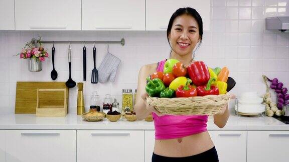 年轻的亚洲运动女性在厨房里拿着一篮子混合蔬菜准备做沙拉看着摄像机健康的食物概念