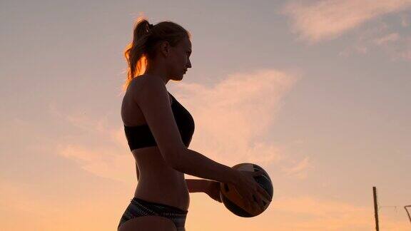 一个美丽的女人穿着比基尼与一个球在日落是准备做发球跳在沙滩上的沙滩排球比赛