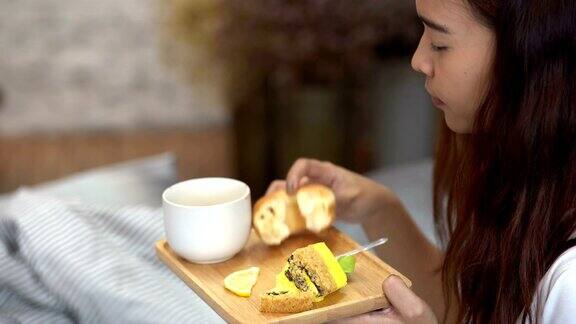 快乐的亚洲女性早上在卧室里吃着蛋糕、甜甜圈和一杯咖啡