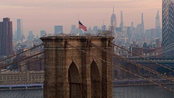 在清晨的黎明时分俯瞰布鲁克林大桥塔顶飘扬的美国国旗遥望曼哈顿无人机视频与静态摄像机