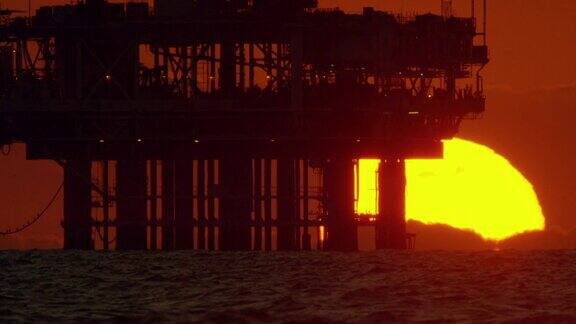 日落在地平线上的慢动作拍摄与一个轮廓的近海石油钻井平台附近亨廷顿海滩在南加州和海浪在前景