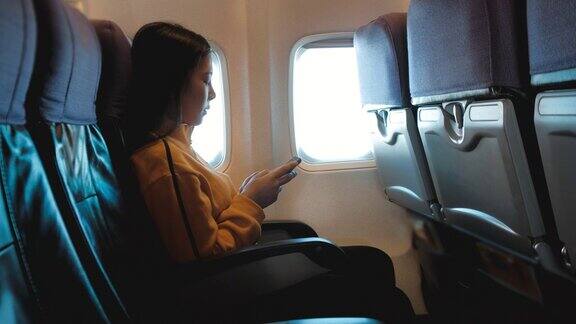 亚洲女游客坐在飞机窗口附近在飞行中使用智能手机