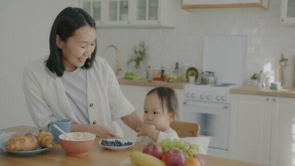 可爱的孩子吃着新鲜的浆果而亚洲妈妈和孩子说话在厨房笑