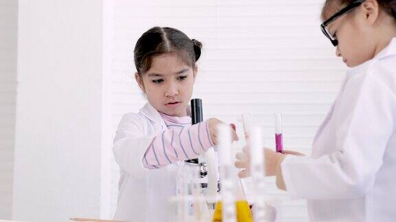 两个小学女生穿着白大褂在科学实验室教室里做实验用试管混合溶液用显微镜分析结果并做笔记