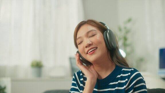 年轻漂亮的亚洲女人戴着无线耳机开心地听着播客音乐唱着歌女人在客厅的沙发上放松