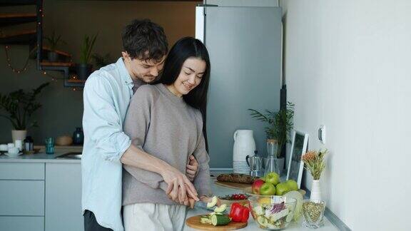 年轻女子切蔬菜烹饪沙拉然后亲吻和拥抱丈夫在厨房的公寓