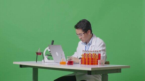 在绿屏背景的实验室里亚洲男性科学家一边在桌子上用显微镜和试管打字一边在笔记本电脑上打字