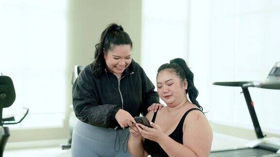 两名肥胖的亚洲女性穿着运动服坐在村俱乐部健身房用智能手机寻找运动后吃饭的餐厅生活方式与运动