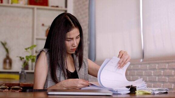 肖像亚洲女人正忙着搜索翻找成堆的文件躺在桌子上头发凌乱卷曲忙着在笔记本电脑屏幕上工作眉头紧蹙在面包店咖啡馆