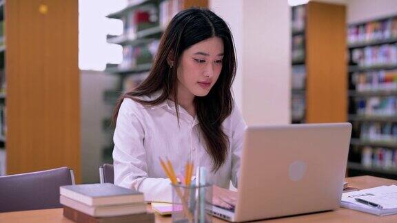 身着便服的亚洲女学生正在大学图书馆用笔记本电脑为论文做笔记