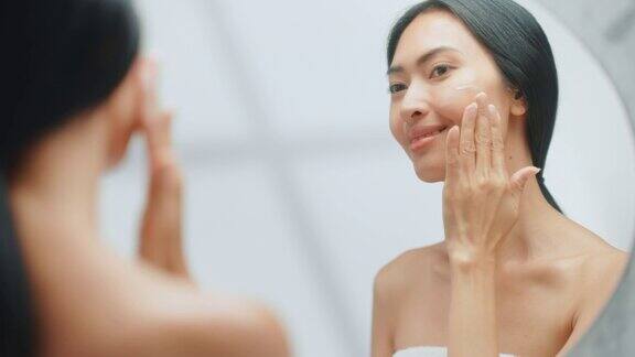 美丽的亚洲妇女肖像轻轻应用面霜面膜感性的触摸看在浴室的镜子年轻女性使她的皮肤柔软清洁与天然化妆品护肤品