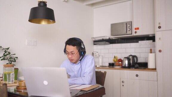 亚洲华人中年男子面带微笑戴着耳机在用餐区使用笔记本电脑参加在线会议