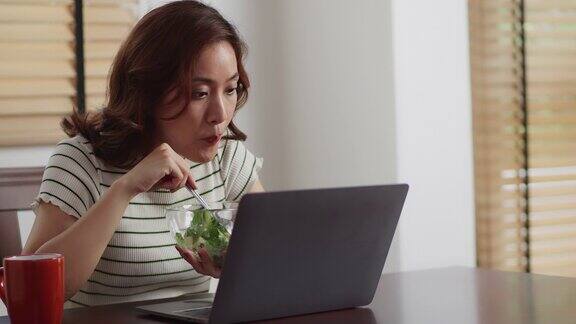 亚洲年轻女子看着笔记本电脑快乐地在家里吃着新鲜的沙拉