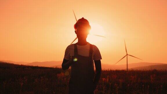 剪影非洲人站在田中与巨大的风车在夏季日落专业技术人员穿着工作服戴橙色头盔和安全眼镜一名风力涡轮机工程师的肖像