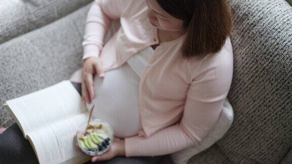 孕妇吃着简单的早餐早上在家里的沙发上读着一本书