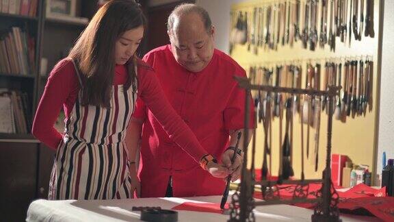 一位亚洲华人父亲在练习中国新年的书法并在一张红纸上教他的女儿握着她的手